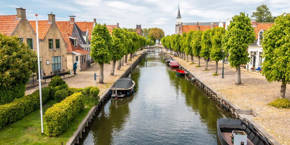 Kanal mit Booten darauf in den Niederlanden (Sloten). Auf beiden Seiten des Kanals sind Bäume und Häuser Allee-artig aufgebaut.