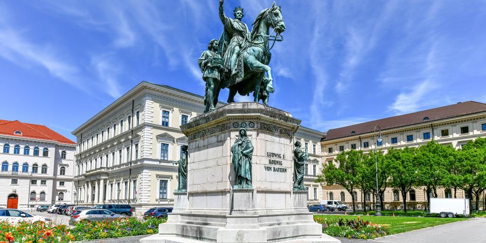 König Ludwig Denkmal in Bayern vor weißen Häuserfassaden und blauem Himmel mit leichten Wolken.