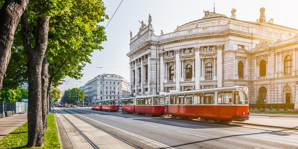 Vor dem Wiener Burgtheater fährt eine weiß-rote Tram auf der Ringstraße. Im Bild sind grüne Bäume, die Sonne scheint. 