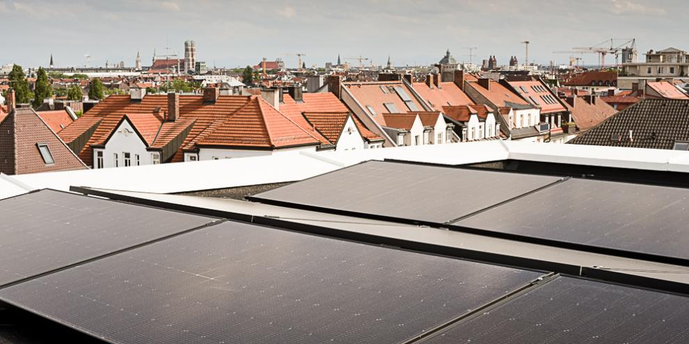 Auf dem Dach des Münchner Verlages sind fünf Solarpanele zu sehen. Im Hintergrund weitere Gebäude und in der Ferne die Münchner Innenstadt und Himmel. 