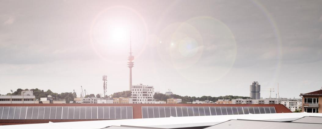 Auf dem Dach des Münchner Verlages scheint die Sonne auf die dort verbauten Solarpanele. Im Hintergrund Himmel, der Olympiaturm und Gebäude.