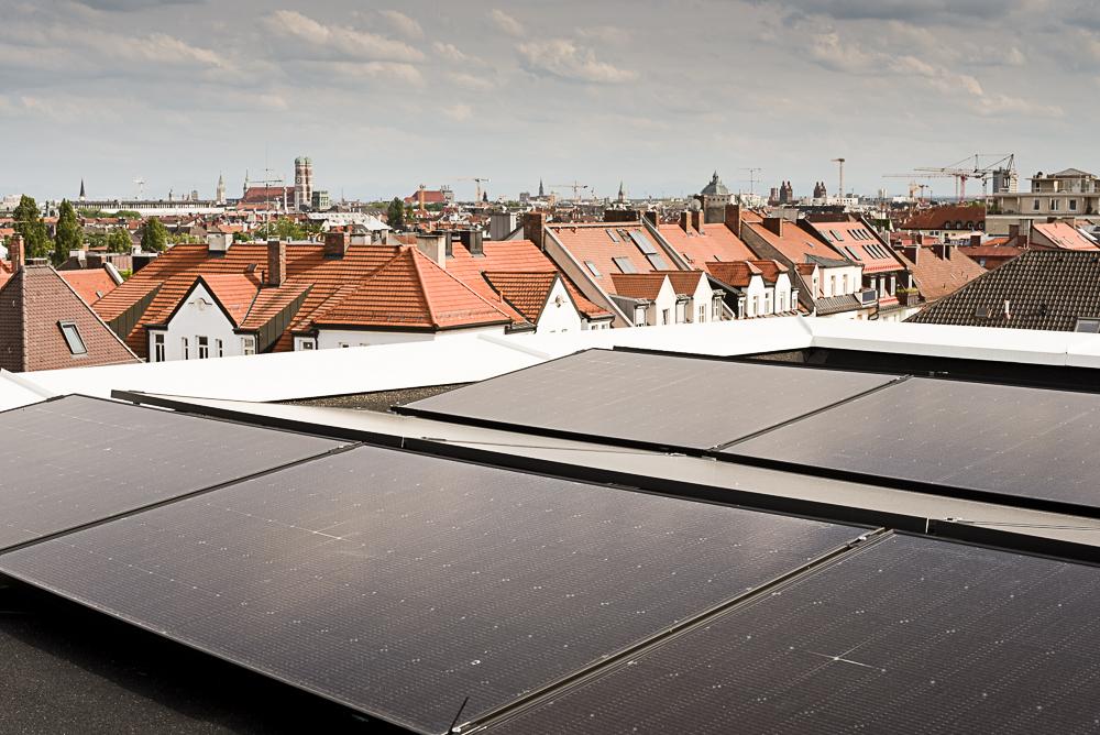 Auf dem Dach des Münchner Verlages sind fünf Solarpanele zu sehen. Im Hintergrund weitere Gebäude und in der Ferne die Münchner Innenstadt und Himmel. 