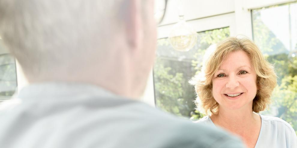 In einem hellen Raum stehen sich zwei Personen gegenüber. Die Frau ist von vorne zu sehen und lächelt den Mann an. 