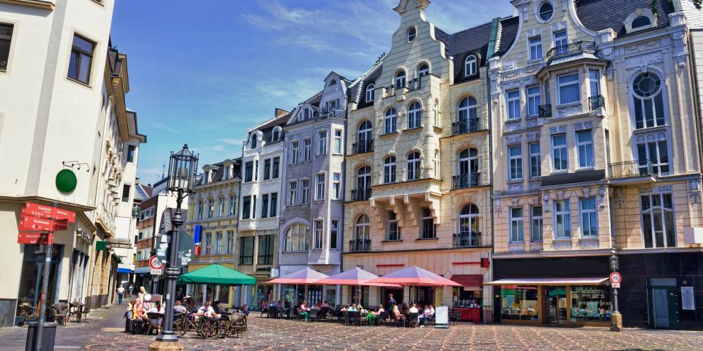 Ein Platz in einer Innenstadt mit Marktschirmen, Ständen und hohen Altbaufassaden bei blauem Himmel. 