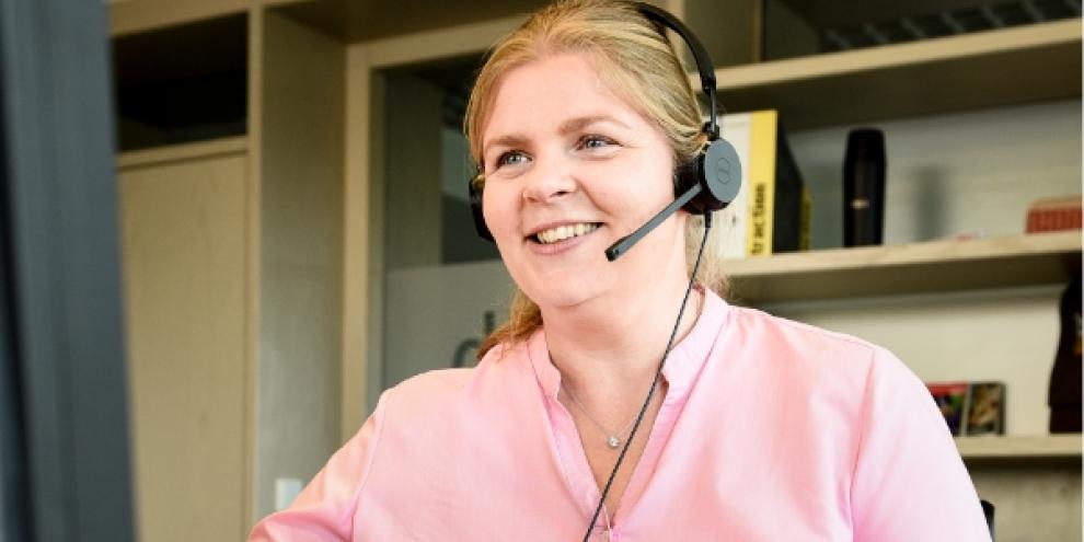 Vor einem Regal sitzt eine Frau, die über ein Headset telefoniert. Sie schaut dabei auf einen Bildschirm. 