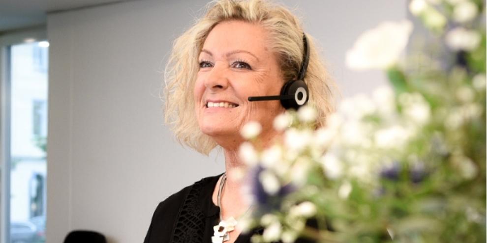 In einem Raum sitzt eine Frau, die ein Headset trägt. Verschwommen im Vordergrund befindet sich ein Blumenstrauß. 