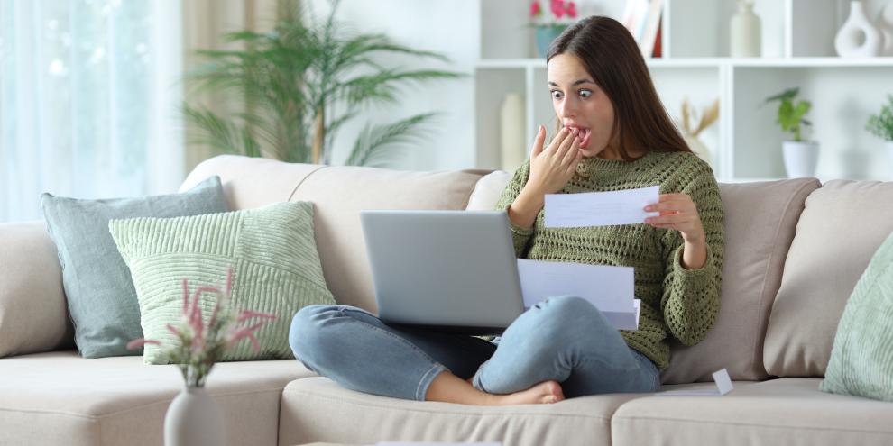 Eine Frau sitzt auf dem Sofa mit dem Laptop auf ihrem Schoß und guckt dabei positiv überrascht.