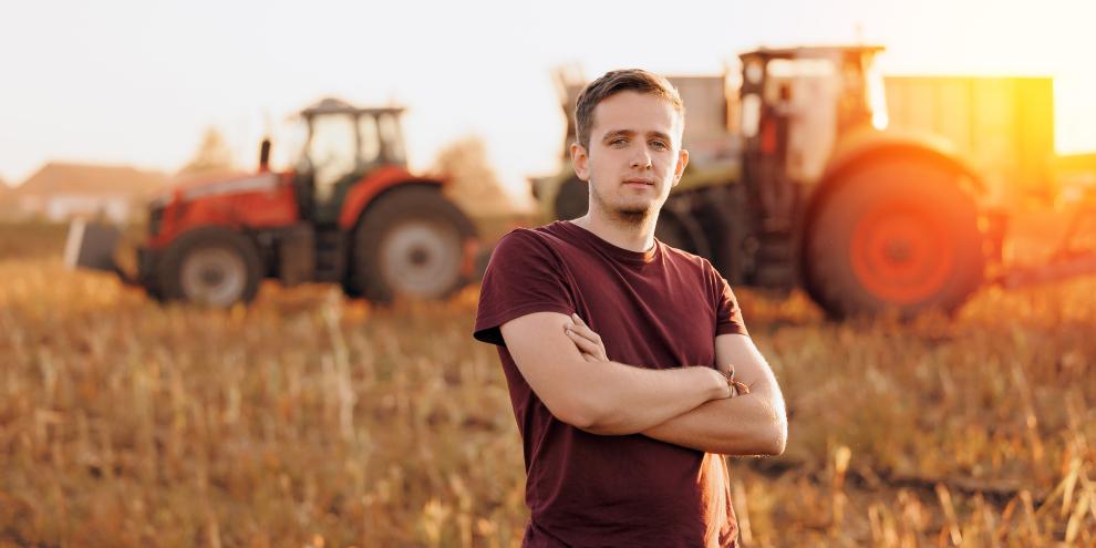 Porträt eines jungen Landwirts in einem Maisfeld, wo im Hintergrund ein Mähdrescher und ein Traktor auf dem abgeernteten Maisfeld zu sehen sind