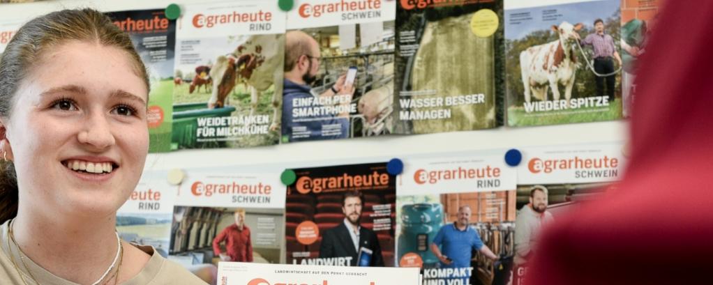 Vor einem Regal mit vielen verschiedenen Zeitschriften sitzt eine junge Frau und lächelt die zwei ihr gegenüber sitzenden Personen an.