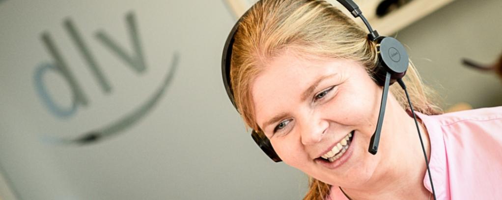 Vor einer Glastür, auf dem das dlv-Logo kaschiert ist, sitzt eine Frau und telefoniert über ein Headset.  