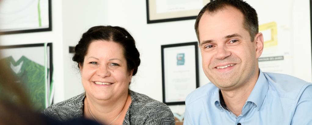 Vor einer Wand mit Bildern und Awards sind zwei Kollegen von vorne und eine Person von der Seite in einem Gespräch zu sehen. 