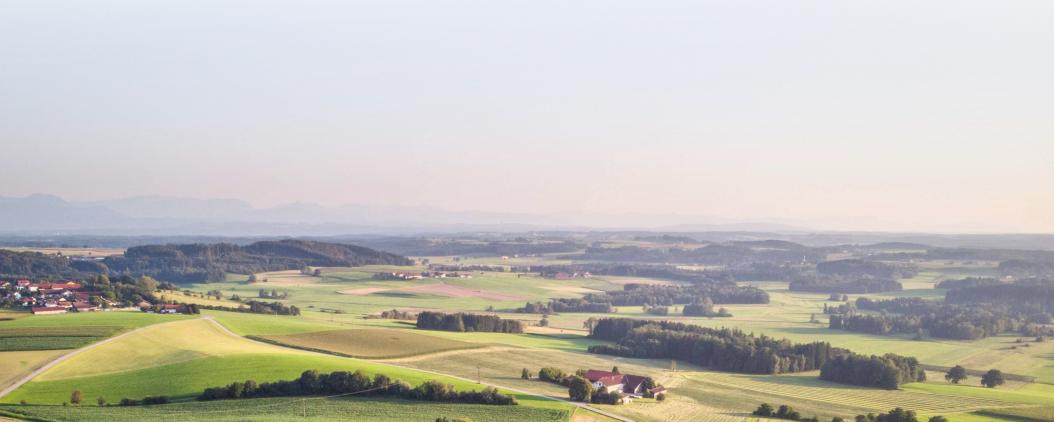 Ein Landschaftspanorama mit Wiesen, Felder und Wäldern, sowie vereinzelten Häusern und Dörfern von oben. Im Hintergrund sind Gebirgszüge. 