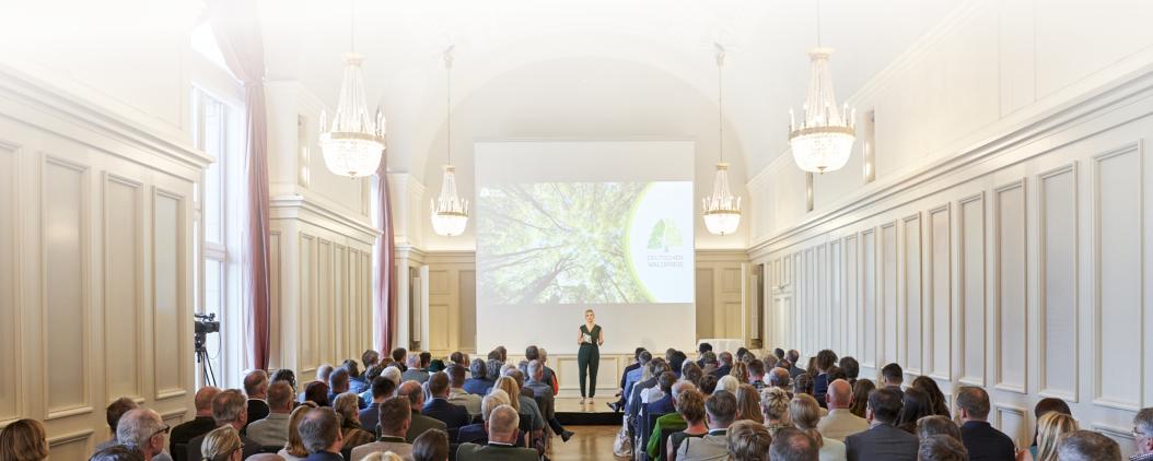 Der Kaisersaal der Deutschen Parlamentarischen Gesellschaft in Berlin ist hell erleuchtet und hat hohe decken. Auf einer Bühne steht eine Frau vor einer großen Leinwand. Im Publikum sitzen viele Menschen in festlicher Kleidung. 