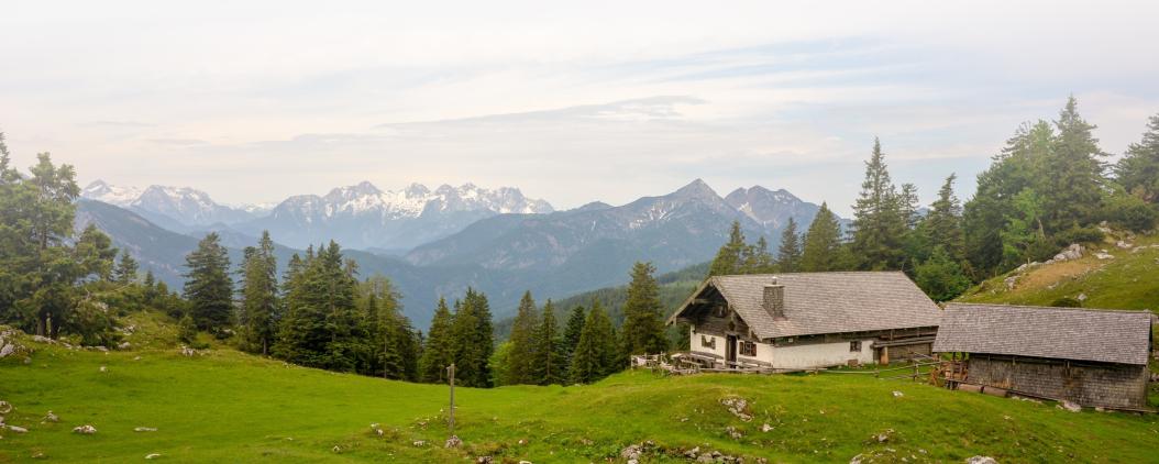 Auf einem felsigen Berg stehen zwei kleine Hütten auf einer Wiese. Im Hintergrund liegen Tannen und schneebedeckte Berggipfel.