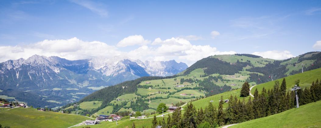 Zu sehen ist ein Bergpanorama, mit schneebedeckten Gipfeln im Hintergrund und grün-bewaldeten Gipfeln im Vordergrund. Dazwischen liegen kleine Dörfer und Liftanalagen. 