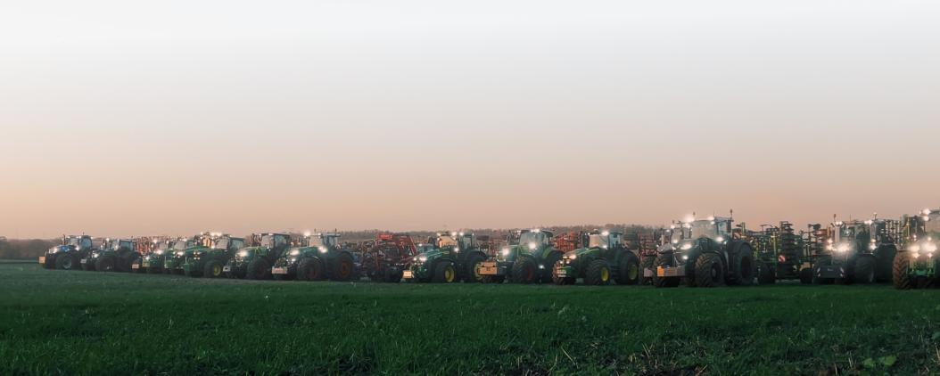 Auf einem grünen Acker stehen viele Trecker aufgereiht und mit Bodenbearbeitungsmaschinen. Es dämmert schon und die Trecker haben Lichter an. 