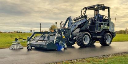 Elektrischer Hoflader mit einem angebauten Kehrbesen fährt auf einer Straße.