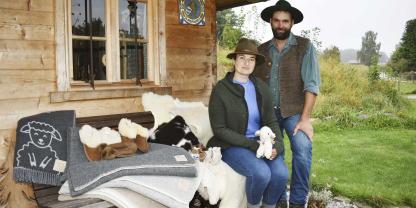 Eine Frau und ein Mann neben Wollprodukten vor einer Hütte.