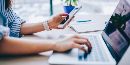 Nahaufnahme einer Frau, die ein Smartphone hält und parallel auf ihrem Laptop tippt
