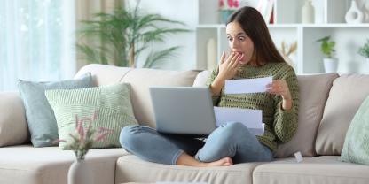 Eine Frau sitzt auf dem Sofa mit dem Laptop auf ihrem Schoß und guckt dabei positiv überrascht.