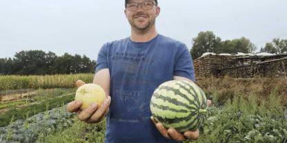 Ein Mann steht auf dem Acker, in jeder Hand hält er eine Melone vor sich. 