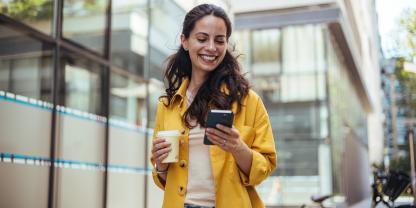 Eine junge Frau geht eine Straße in der Stadt entlang, schreibt Nachrichten und hält dabei einen Kaffeebecher in der Hand.